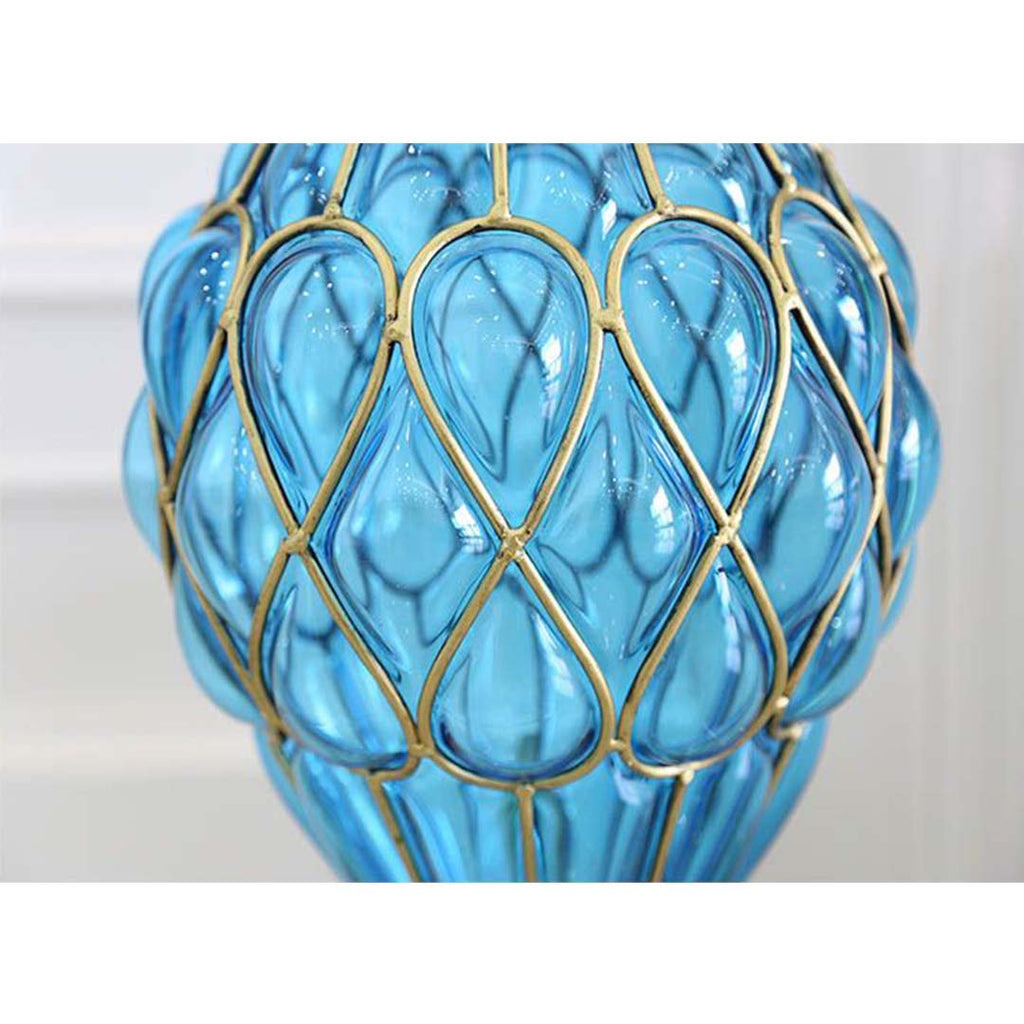 Close-up of a decorative blue glass object with teardrop-shaped forms outlined in gold metal, creating a repeating pattern around its bulbous surface; the craftsmanship and material contrast suggest it may be part of a lamp, vase, or ornamental sculpture.