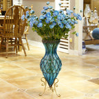 Decorative blue glass vase with intricate swirling patterns, placed on a gold metal stand with three scrollwork legs; filled with vibrant blue artificial flowers and green foliage, set in a tiled room with wooden dining furniture and warm ambient decor.